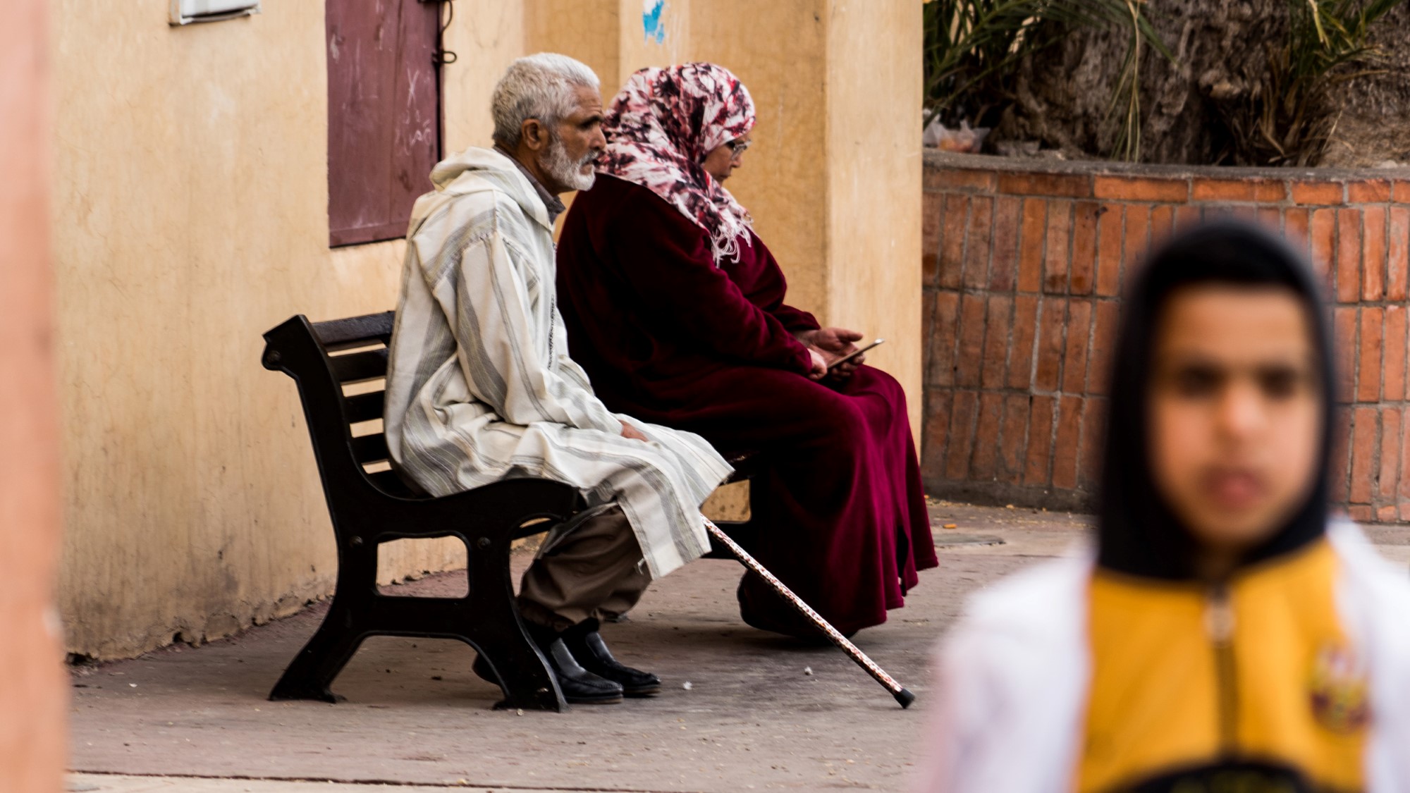 Eldre mann med krykke og eldre kvinne på benk. Ung kvinne i forgrunnen