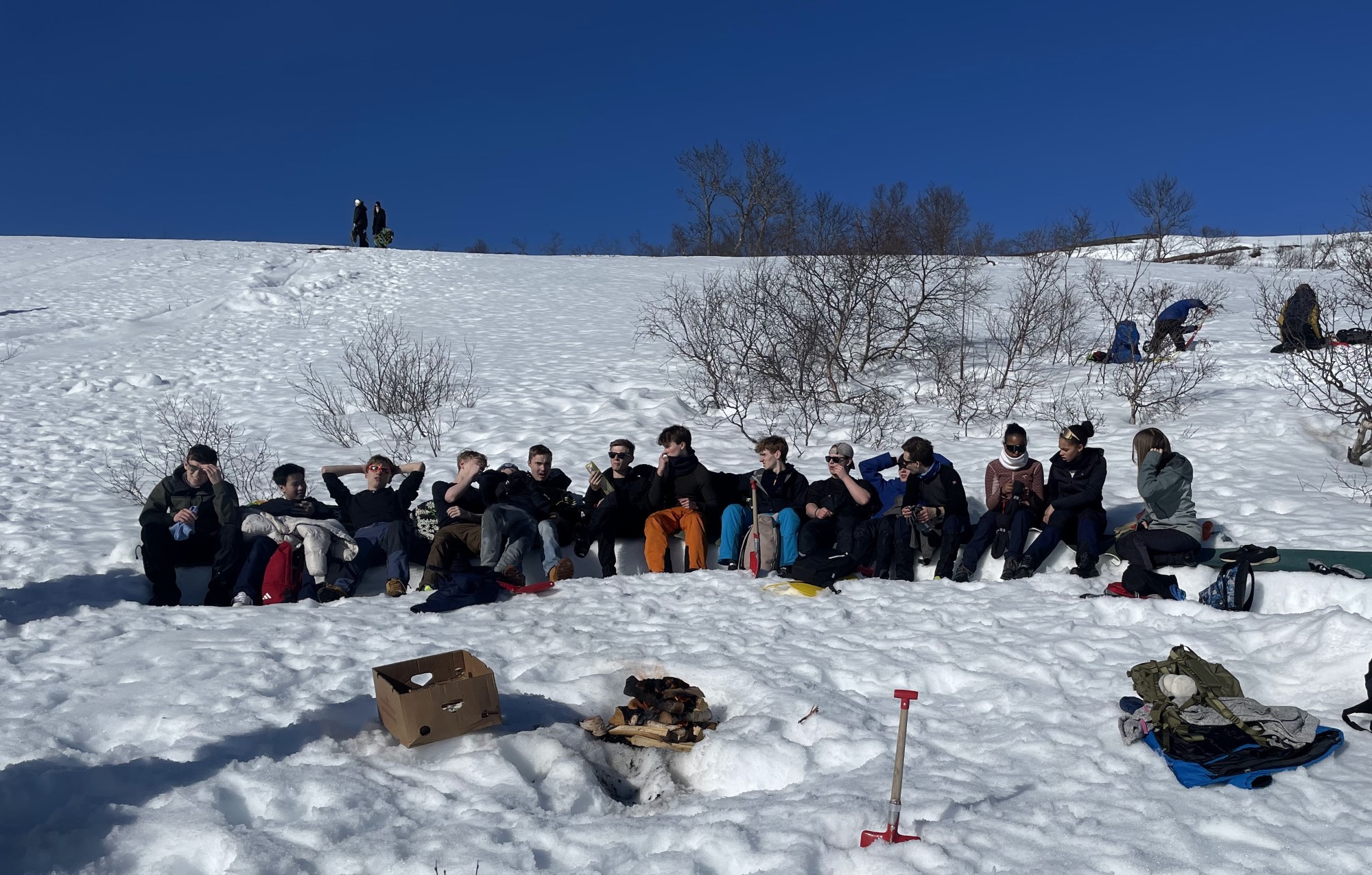 Eng gjeng ungdommer slapper av på påskefjellet foran et bål. Omgitt av blå himmel, sol og snø