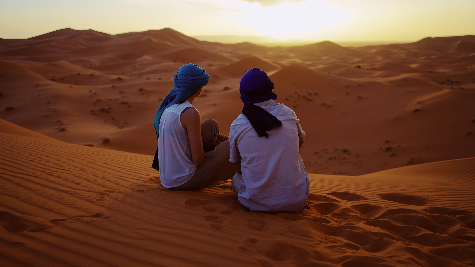 Mann og kvinne sitter i ørkenen i solnedgang med tørkler på hodet.